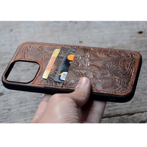 May include: A brown leather phone case with a floral embossed design. The case has a built-in card holder with two cards inserted.