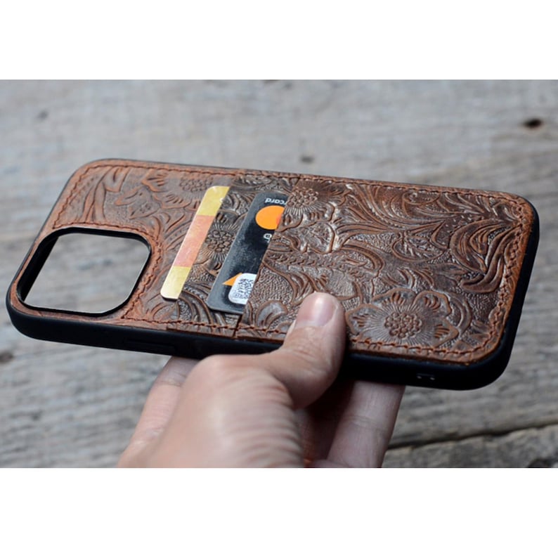 May include: A brown leather phone case with a floral embossed design. The case has a built-in card holder with two cards inserted.