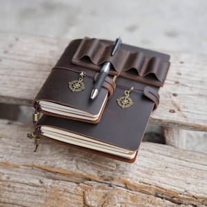 Può includere: Due diari in pelle marrone impilati, con un portapenne e penne. Ogni diario ha un ciondolo a forma di bussola. I diari sono rilegati con un cinturino in pelle e sono su una superficie di legno.