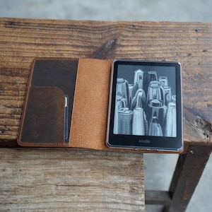 Könnte beinhalten: Geöffnete Kindle-Hülle aus braunem Leder mit Kartenfach. Ein Kindle-Gerät mit Schwarz-Weiß-Bild ist zu sehen. Die Hülle liegt offen auf einer Holzoberfläche.