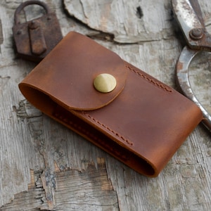 May include: A rectangular brown leather pouch featuring a rounded flap secured by a single brass-colored metal snap button. The leather has a matte, textured finish with visible stitching along the edges, creating a simple, functional storage case.