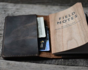 May include: A brown leather notebook cover with a Field Notes notebook inside. The notebook cover has a pocket for cards and cash. The notebook is open to reveal the wood cover and the text "FIELD NOTES" printed on the front.
