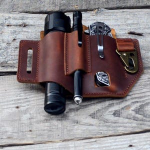 Puede incluir: Una funda de cinturón de cuero marrón con varios bolsillos para llevar una linterna, un bolígrafo y una navaja plegable. La funda tiene un mosquetón de metal.
