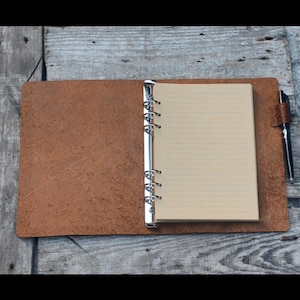 May include: A brown leather bound notebook with a silver ring binder and lined paper. The notebook is open and lying on a wooden surface.