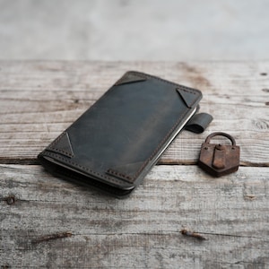 May include: A dark gray leather phone case with brown stitching and corner accents. A small, rusty padlock sits next to the case on a weathered wooden surface. The case has a magnetic closure.