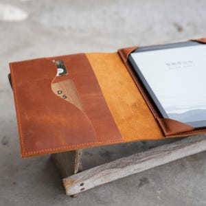 May include: Open, brown leather tablet case with a card slot and a tablet inside. The case is made of a smooth, textured leather and has a contrasting lighter brown interior. The tablet screen displays text in a foreign language.