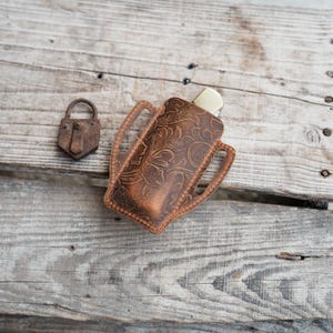 Pode incluir: Uma bainha de faca de couro castanho com um design floral em relevo. A bainha tem uma pega e contém uma faca com uma pega creme. Um pequeno cadeado enferrujado está à esquerda da bainha.