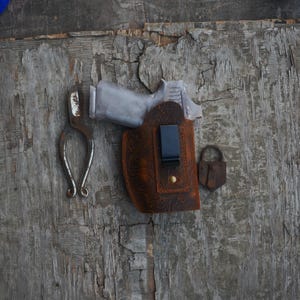 May include: A brown leather holster with a floral design, a silver gun, and a small silver tool on a wooden surface.