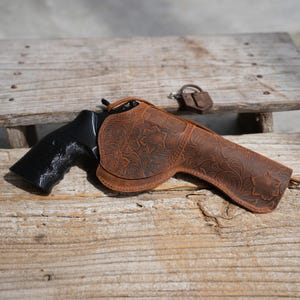 Peut inclure: Un étui en cuir marron avec des motifs floraux, conçu pour un revolver. L'étui est posé sur une surface en bois vieilli, avec un revolver noir et un petit accessoire en cuir. L'étui présente un motif floral décoratif.