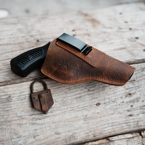 Op de afbeelding: Een bruin leren holster met een bloemmotief en een zwarte metalen clip. Een zwarte pistoolgreep is zichtbaar naast het holster, samen met een klein, roestig hangslot. De items liggen op een verweerd houten oppervlak.