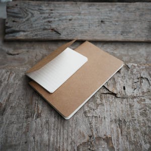 May include: Two brown notebooks, one with a partially opened, lined white page, rest on a weathered wooden surface. The notebooks are rectangular with rounded corners. The wood has a rustic, aged appearance.