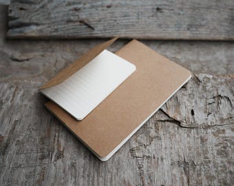 May include: Two brown notebooks, one with a partially opened, lined white page, rest on a weathered wooden surface. The notebooks are rectangular with rounded corners. The wood has a rustic, aged appearance.