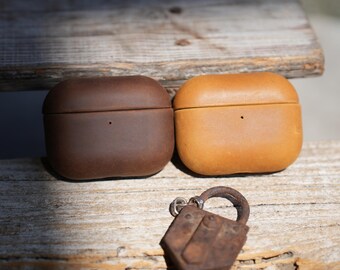 May include: Two leather AirPod cases, one dark brown and one tan, sit side-by-side on a weathered wooden surface. A small, rusty metal padlock with a leather loop hangs below the cases. The cases have a smooth, rounded design.