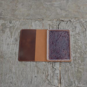 Puede incluir: Un cuaderno de cuero marr&oacute;n con un inserto de pl&aacute;stico transparente que contiene una almohadilla de tinta morada.