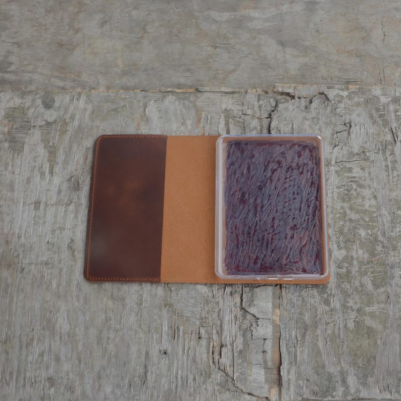 Puede incluir: Un cuaderno de cuero marr&oacute;n con un inserto de pl&aacute;stico transparente que contiene una almohadilla de tinta morada.