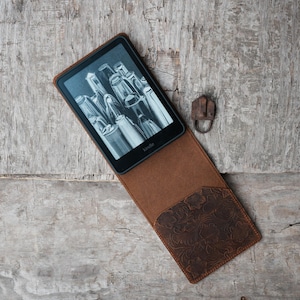 Puede incluir: Una funda Kindle de cuero marrón con un diseño floral en relieve en el bolsillo. El Kindle muestra una imagen en blanco y negro. La funda está abierta y el Kindle es visible. El fondo es una superficie de madera envejecida.