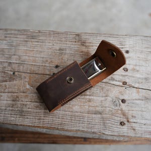May include: A dark brown leather case with visible stitching and a metallic snap button closure, containing a silver-coloured harmonica. The case is constructed from thick, matte-finished leather and features a flap that secures the instrument inside.