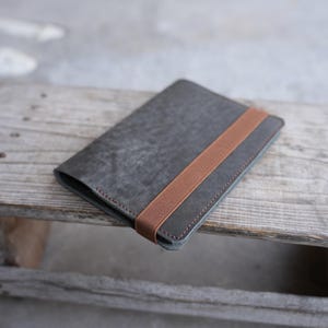 May include: A gray leather wallet with a brown elastic band closure. The wallet is rectangular and has visible stitching around the edges. The leather has a textured appearance, and the wallet is resting on a wooden surface.