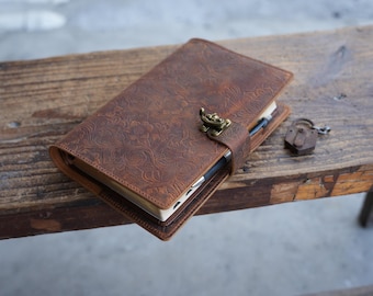 Könnte beinhalten: Ein braunes Lederjournal mit geprägtem Blumenmuster. Das Journal ist mit einem Messingverschluss verschlossen und hat ein passendes kleines Schloss. Das Journal liegt auf einer Holzoberfläche.
