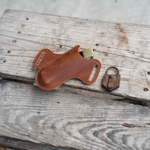 Puede incluir: Una funda de cuero marrón para cuchillo con un cuchillo de color latón parcialmente insertado. Junto a ella, un pequeño candado oxidado. Los objetos están colocados sobre una superficie de madera envejecida, sugiriendo un tema rústico o al aire libre.