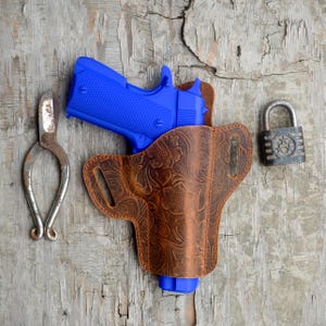 Puede incluir: Una pistola azul en una funda de cuero marrón con un diseño floral. También hay un candado plateado en la imagen.