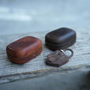 May include: Two leather cases in shades of brown, alongside a small, aged metal padlock. The cases are rectangular with curved sides. The padlock has a ring. The items are on a wooden surface.