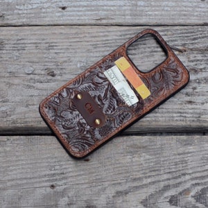 May include: A brown leather phone case with a floral embossed design. The case has a built-in card holder and a metal plate with the initials "CM" engraved on it.