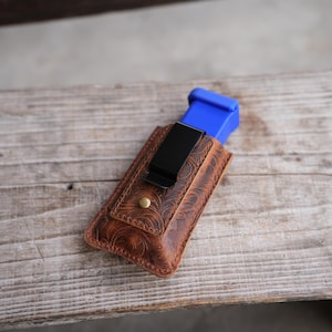 May include: A brown leather holster with an intricate floral embossed pattern, featuring a black metal belt clip, a single brass-colored rivet, and a bright blue rectangular object inserted into the top opening of the leather pouch.