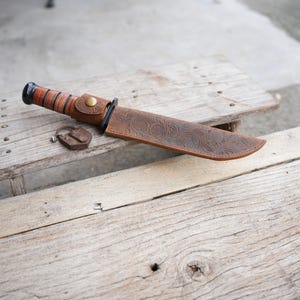 May include: A large, brown knife with a leather sheath and handle. The sheath features an embossed floral pattern and a brass button closure. A small, rusty padlock sits next to the knife on a wooden surface.