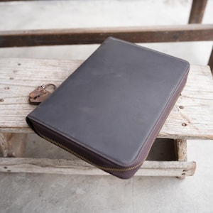 May include: A dark grey leather portfolio with a brass zip closure. The rectangular portfolio is closed. An antique metal lock sits on the wooden surface beside the portfolio. The portfolio is likely for documents or a tablet.