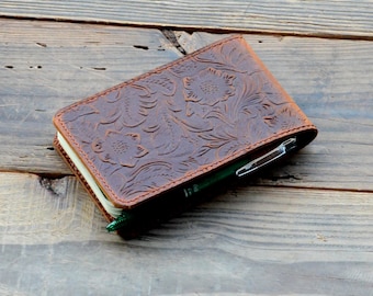 May include: A brown leather notebook cover with an embossed floral design. The cover has a pen holder and is closed.