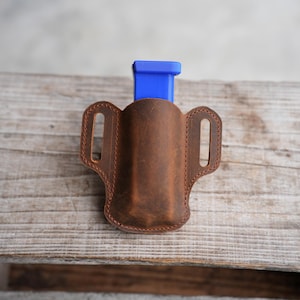 Puede incluir: Una funda de cuero marrón para cargador con una ranura central y dos trabillas laterales para el cinturón. Un cargador de plástico azul brillante está insertado en la parte superior de la funda, que presenta costuras marrones visibles en los bordes.