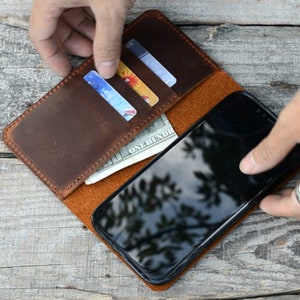 May include: A brown leather phone case with a flap that opens to reveal slots for credit cards and cash. The case is open and a black smartphone is visible inside.