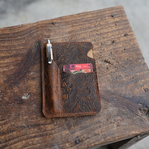 May include: A brown leather pocket organizer featuring an embossed floral pattern. The organizer includes a dedicated vertical slot holding a silver and wood pen, and a separate pocket containing a rectangular card with red and white text.