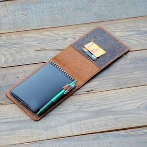May include: A brown leather notebook cover with a floral embossed design. The cover has a pocket for cards and a pen holder. A black notebook and a green pen are inside the cover.