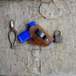 May include: A blue and brown leather gun holster with a black clip and a padlock on a wooden surface.