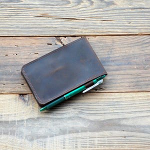 Puede incluir: Una cartera de cuero marrón oscuro con un bolígrafo dentro, sobre una superficie de madera. La cartera tiene un borde cosido y un diseño sencillo. El bolígrafo es verde y blanco.