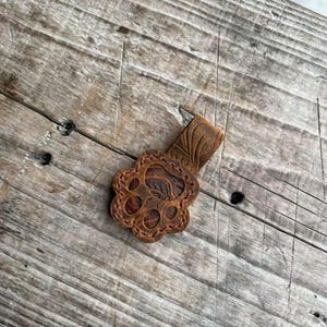 May include: A brown leather keychain with a paw print design. The keychain has a rectangular strap and a paw-shaped charm with detailed paw pad and claw impressions. The leather has a natural, distressed appearance, and the background is a weathered wooden surface.