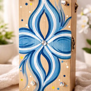 May include: A wooden box with a hand-painted blue and white floral design. The design is centered on the front of the box and features a crystal embellishment in the centre. The box has a light brown finish and is decorated with small dots.