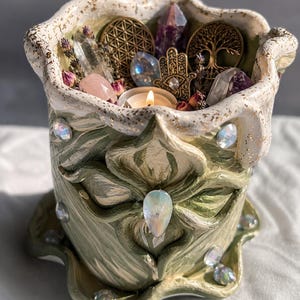 Può includere: Un contenitore in ceramica verde e bianco fatto a mano con un motivo floreale, adornato con cristalli iridescenti. Il contenitore contiene cristalli, fiori secchi, una candela accesa e ciondoli decorativi in metallo. Il bordo ha accenti dorati.