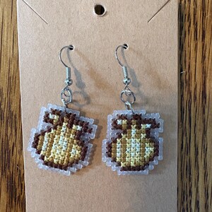 May include: A pair of brown and yellow cross-stitch earrings shaped like money bags hanging on a brown card.