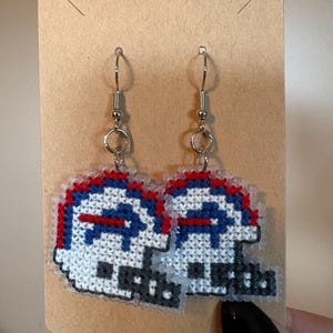 May include: A pair of cross-stitched earrings featuring a football helmet design in white, blue, and red.