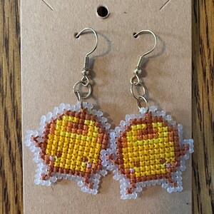 May include: A pair of yellow and brown cross-stitch earrings with gold hooks. The earrings are shaped like a cartoon character.