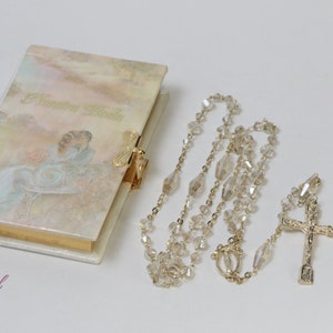 May include: A white book with a gold clasp and a watercolor image of a woman in a white dress. The book is next to a gold chain with clear crystal beads and a gold cross.
