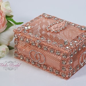 Può includere: Scatola in metallo color oro rosa con un design decorativo di strass trasparenti e un motivo floreale. La scatola ha un coperchio con cerniera ed è decorata con due anelli intrecciati.