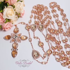Può includere: Rosario color oro rosa con un ciondolo a forma di croce e più fili di perline sfaccettate. La croce e il ciondolo presentano grandi cristalli scintillanti. L'immagine include anche un bouquet di rose rosa e crema. Il testo "Yolis Bridal" è visibile.