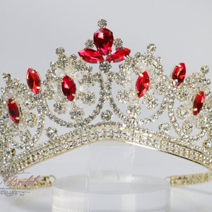 以下が含まれることがあります： 赤と透明なラインストーンが付いた金の王冠。王冠は尖った頂上と湾曲したベースを持っています。