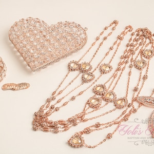 May include: A set of rose gold jewelry featuring a heart-shaped clutch, a small round box, and a multi-strand necklace with a cross pendant. The jewelry is adorned with sparkling crystals.
