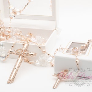 Può includere: Un rosario in oro rosa con perline di cristallo trasparente e un ciondolo a forma di croce, presentato in una scatola bianca aperta. Il rosario presenta un design a croce dettagliato ed è presentato su una superficie bianca. È visibile anche una scatola più piccola con un rosario abbinato.