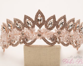 Puede incluir: Una tiara de oro rosa con cristales transparentes y delicados detalles florales. La tiara está sobre un soporte de tela beige.